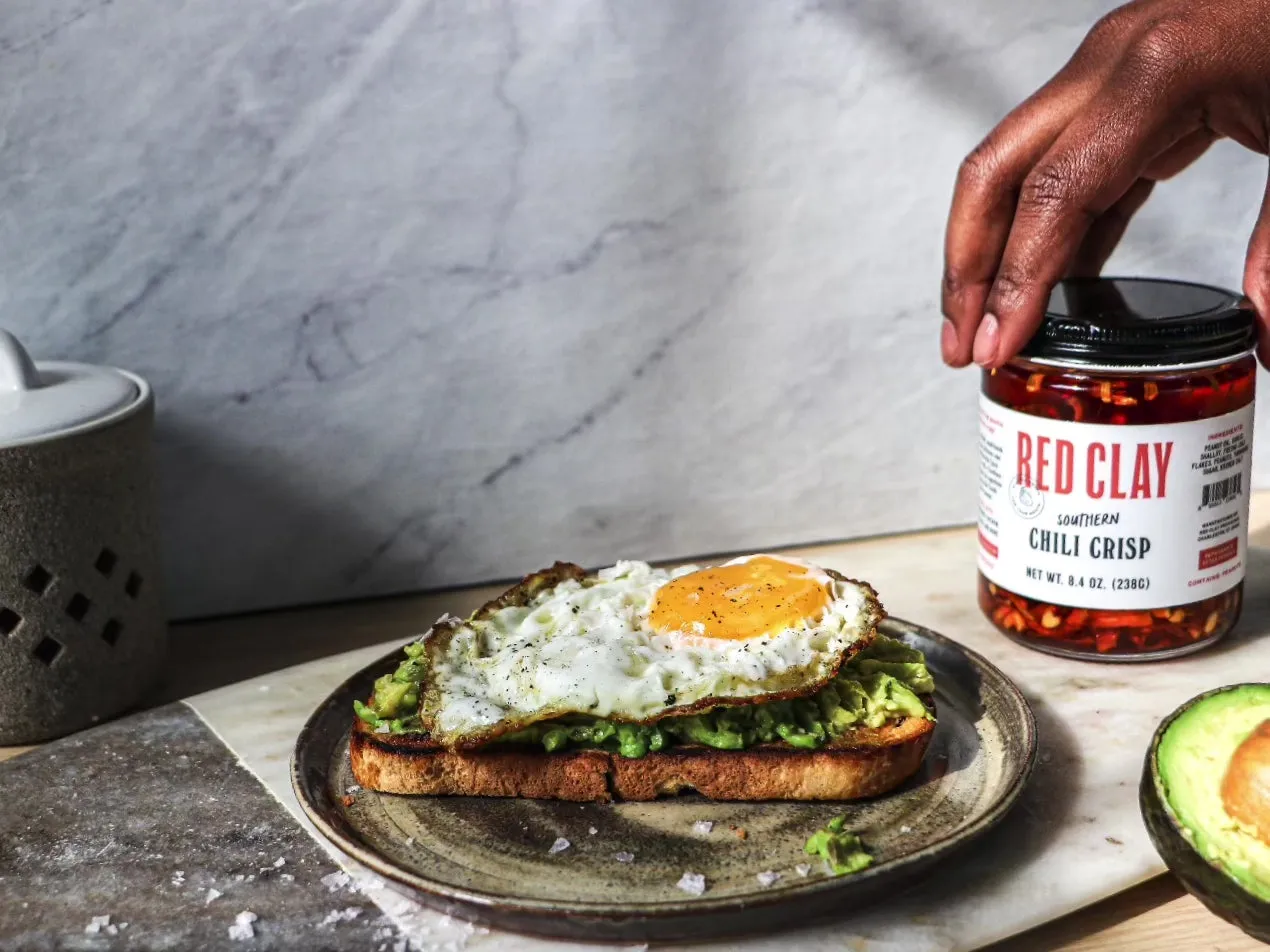 A hand setting a jar of Red Clay Southern Chili Crisp beside avocado toast with a fried egg
